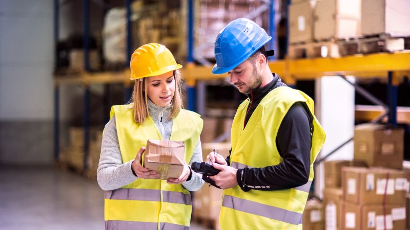 A lady and man wearing high-viz jackets and safety helmets stand in front of a warehouse full of stock as they make notes.