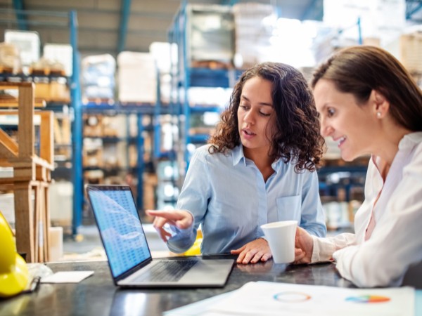 two women sit at a desk with their laptop looking over numbers and doing inventory planning in a manufacturing backdrop.