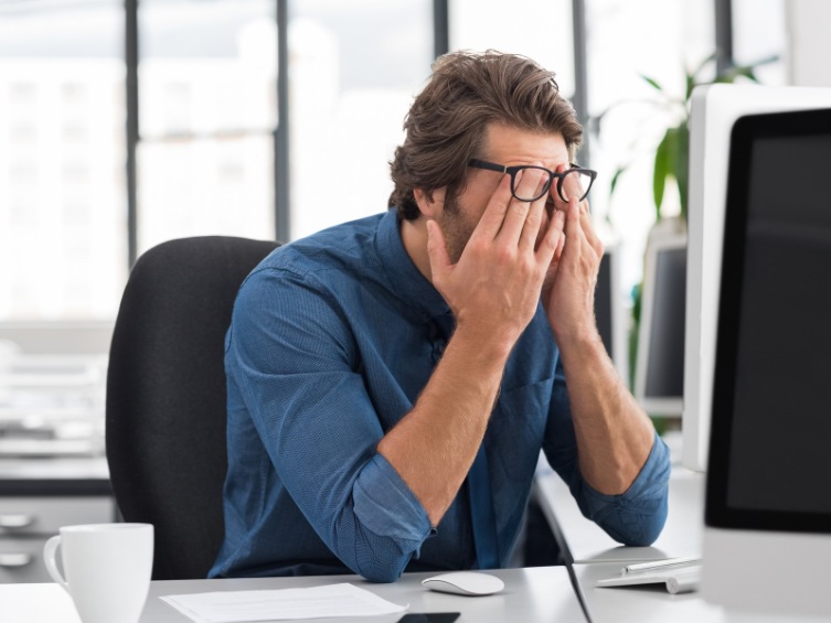 A man in a blue shirt sitting in front of his computer with his head in his hands