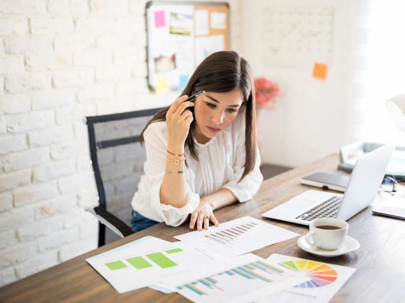 A lady with long dark hair looks confused as she sits at her desk with her laptop and lots of papers with charts and graphs.