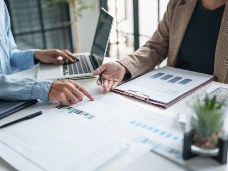 Two people sit at a table with a laptop and a clipboard with bar charts to discuss a business plan for investment