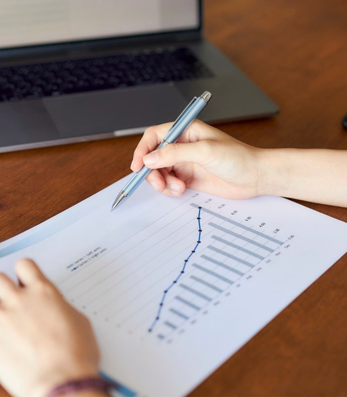 A hand holding a pen hovers a piece of paper showing an increasing graph to indicate financial growth tracking