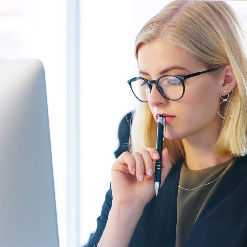 A blonde lady with glasses thinks whilst pressing a pen to her lips and sitting in front of her computer