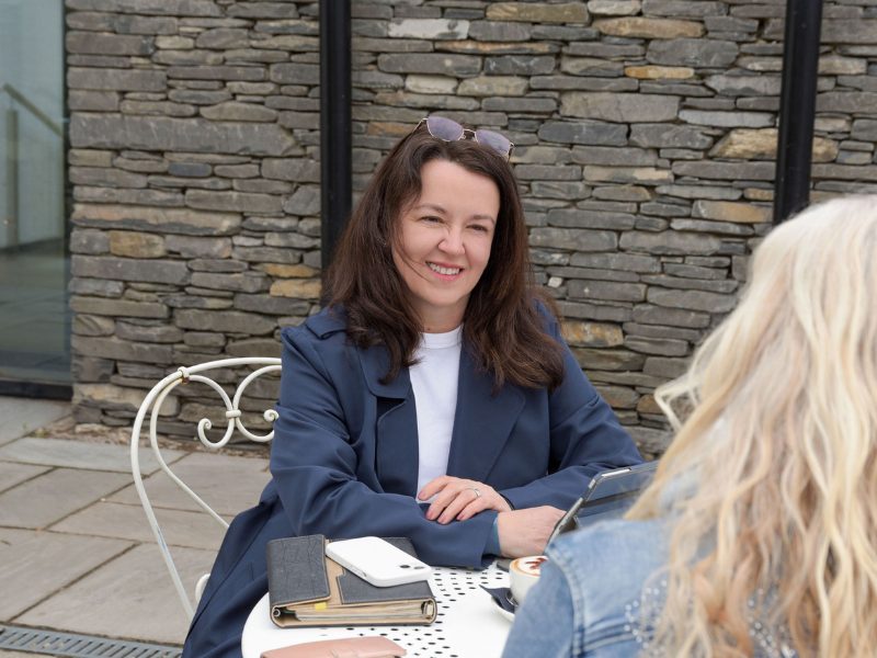 Pauline Healey from Logical BI sitting with a female client outside at a table. She is smiling and wearing a blue coat.
