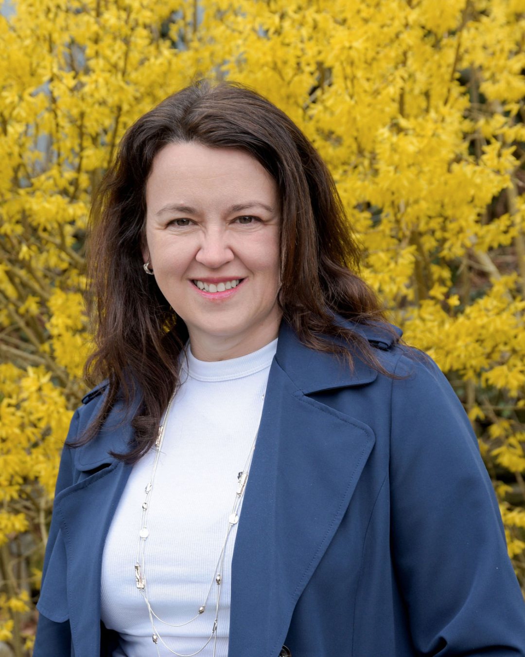 Business Financial Advisor Pauline Healey from Logical BI stands wearing a blue jacket with white t-shirt underneath. She is standing in front of yellow flowers and smiling