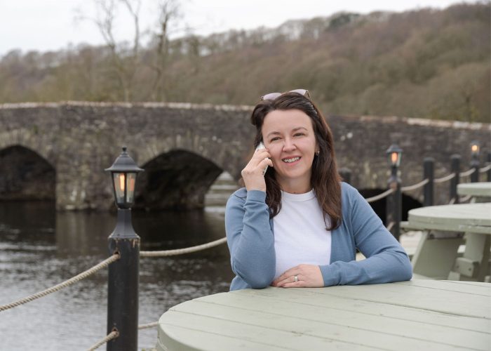 Logical BI founder Pauline Healey sits at a table outside, smiling as she talks on her phone with a river and bridge behind her