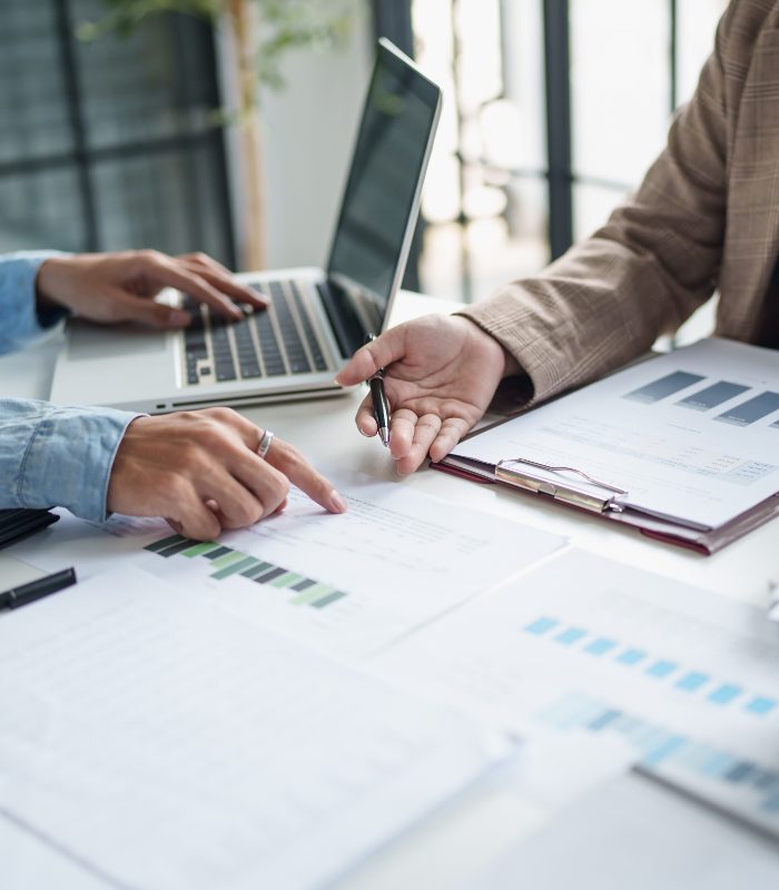2 people sit at a desk opposite each other, gesturing over graphs on paper.