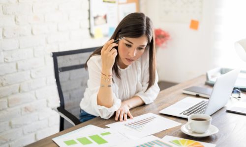 A lady with long dark hair looks confused as she sits at her desk with her laptop and lots of papers with charts and graphs.